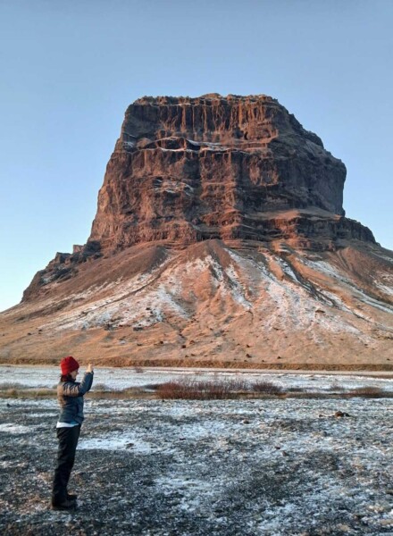 Woman in red beanie and green coat photographing and Icelandic glacier while standing in front of an otherworldly rock formation